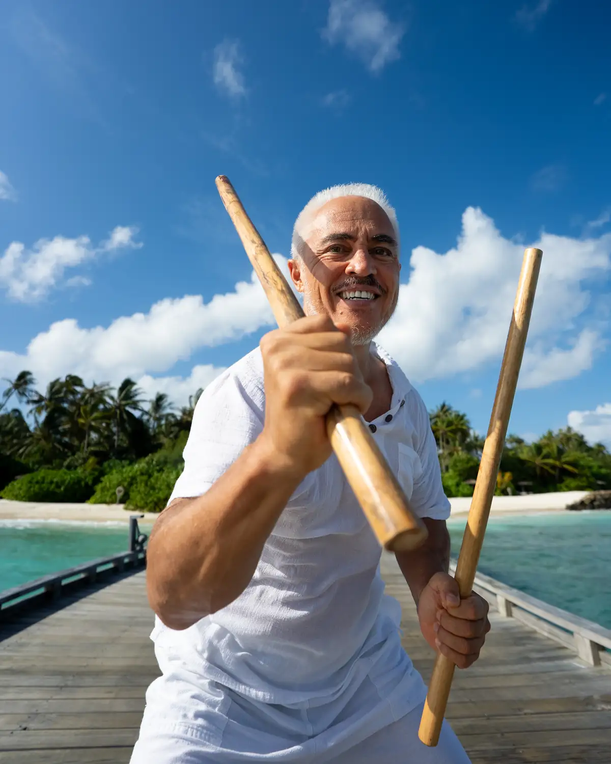 Karol Hasegawa smiling while training with Filipino Kali eskrima sticks on an overwater jetty in the Maldives