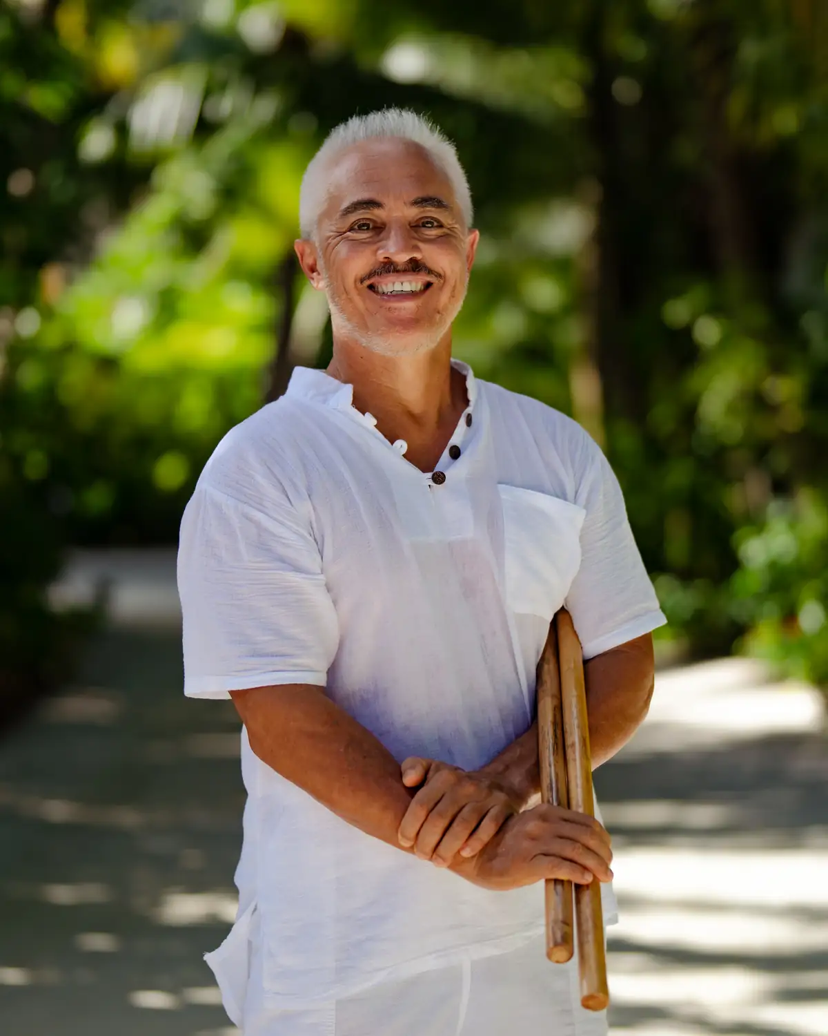 Karol Hasegawa smiling in white attire holding Filipino Kali sticks on a tropical path at JOALI BEING Maldives
